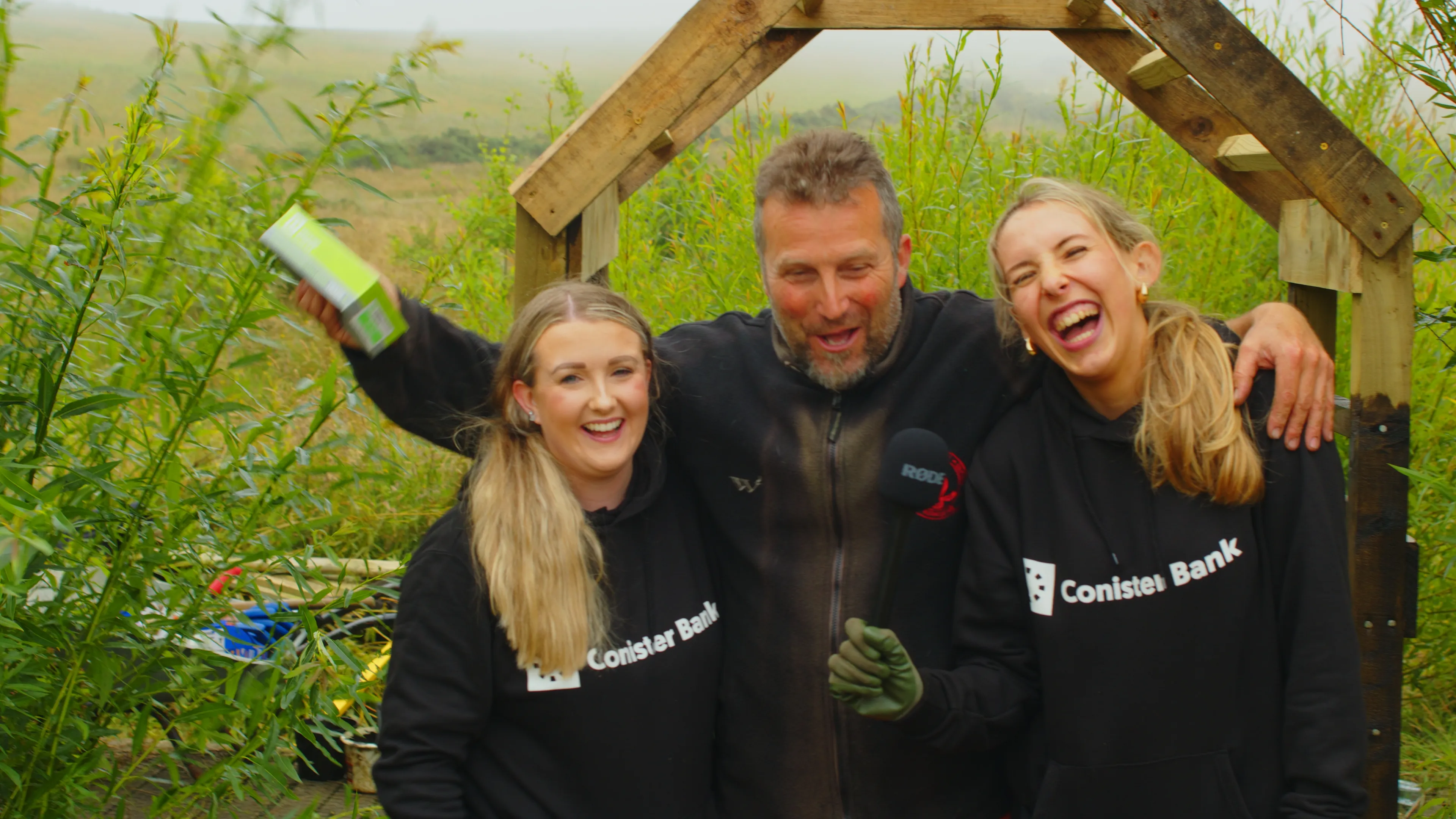 A group of three people laughing in a garden under a wooden archway. Two women in matching black "Conister Bank" hoodies flank a man holding a rectangular box, with greenery in the background.
